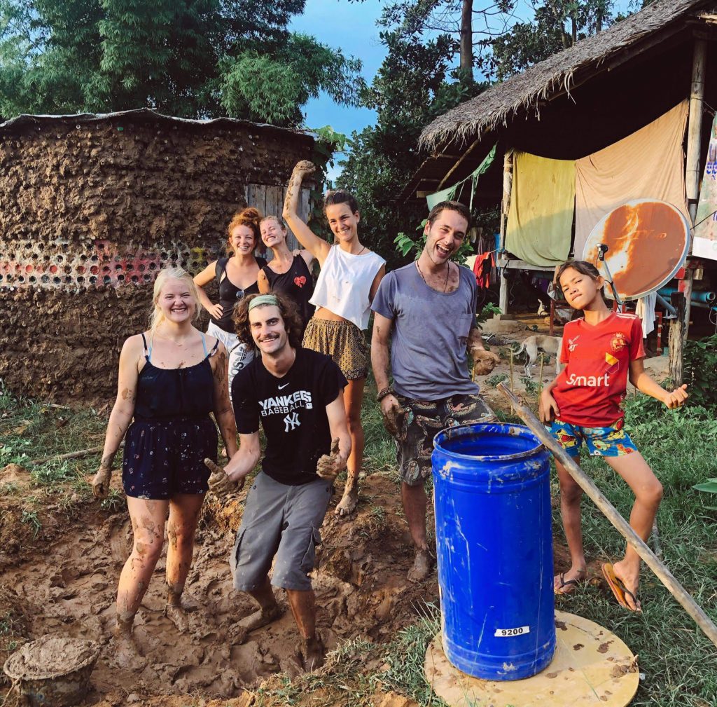 International interns covered in mud smiling with a local child during a natural cob building project in a Cambodian village