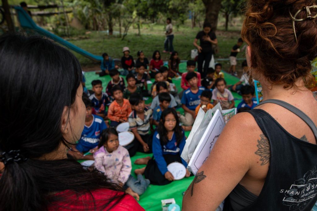 Volunteers leading an outdoor lesson with a large group of Cambodian children seated on a mat at the Red Road Foundation
