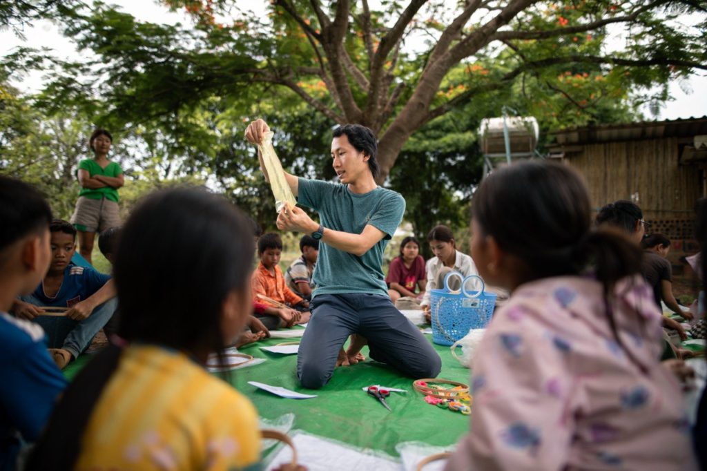 Facilitator leading a hands-on craft lesson with Cambodian children gathered around outdoors at the Red Road Foundation